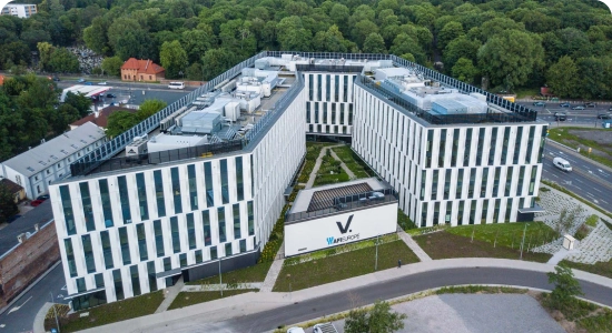 Nowoczesny, biały biurowiec z pionowymi czarnymi pasami i wyposażeniem dachu, otoczony zielenią i drogami, widziany z góry. Z przodu widnieje napis V.AFI EUROPE .