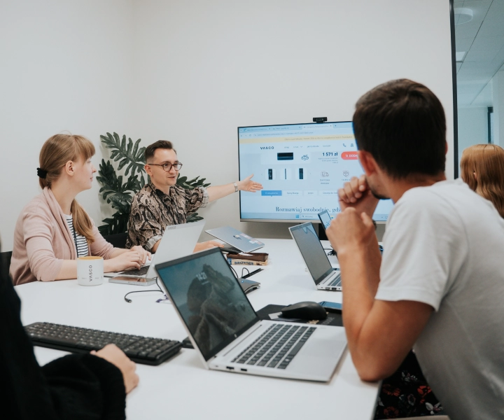 Pięć osób siedzi przy stole z laptopami w sali konferencyjnej. Jedna z osób wskazuje na prezentację na dużym ekranie, na którym wyświetlane są wykresy i zdjęcia produktów. Grupa jest zaangażowana i skupiona na ekranie.