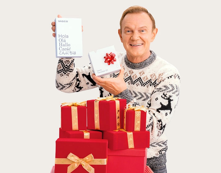 Uśmiechnięty Cezary Pazura w świątecznym swetrze trzyma opakowania produktów Vasco. Wokół niego leżą paczki z prezentami