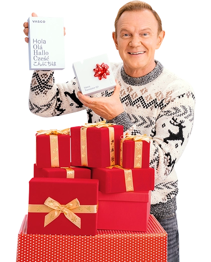 Cezary Pazura stojący za prezentami, w zimowym swetrze, trzyma w rękach pudełko z Vasco Translatorem.