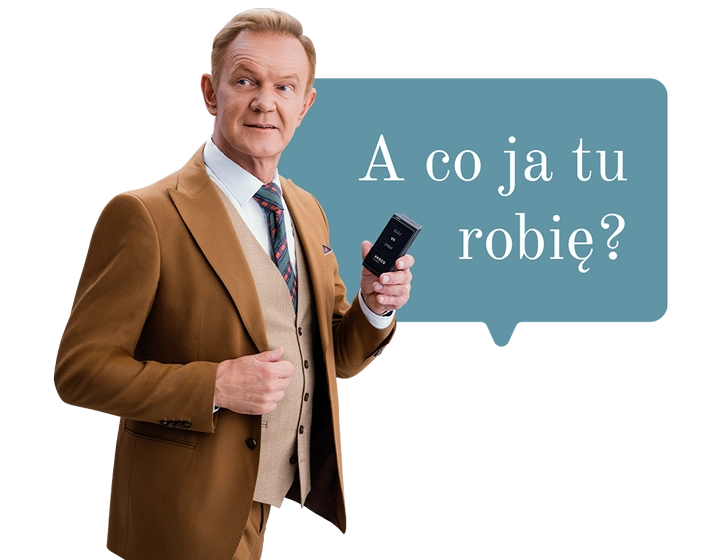 Cezary Pazura w brązowym garniturze, trzymający w ręce Vasco Translator Q1. W tle dymek z tekstem: A co ja tu robię?