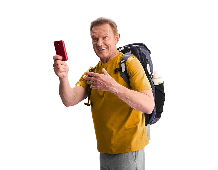 Uśmiechnięty Cezary Pazura ma ręce pełne pudełek z Vasco Translatorami. Ubrany jest w czerwony świąteczny sweter i owinięty lampkami.