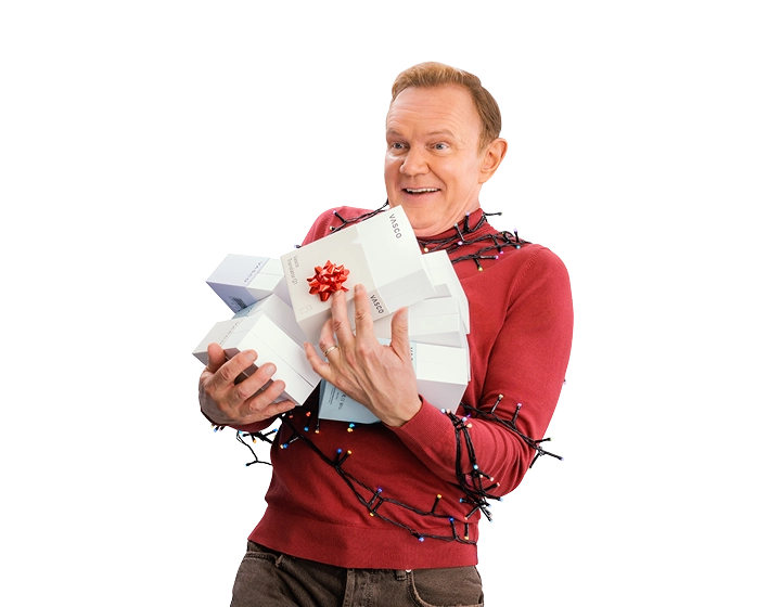 Uśmiechnięty Cezary Pazura ma ręce pełne pudełek z Vasco Translatorami. Ubrany jest w czerwony świąteczny sweter i owinięty lampkami.