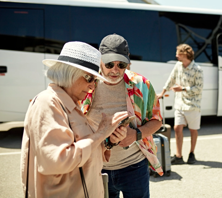 Starsza para patrzy na telefon. W tle widać autobus i osobę z walizką