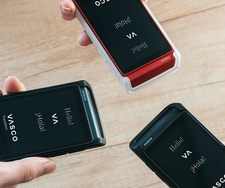 Trzy osoby trzymające urządzenia Vasco Translator Q1 łączą je; na ekranach pojawia się ekran startowy z logo Vasco oraz napisami „Hello!” i „¡Hola!”. Urządzenia są dostępne w silikonowych etui w trzech kolorach: Solid Black, Dark Caramel i Milky White.