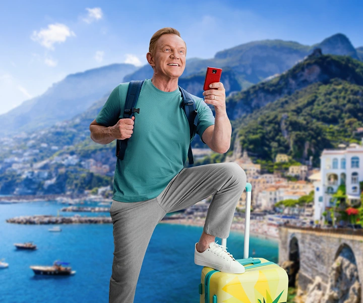 Cezary Pazura z plecakiem opiera nogę na żółtej walizce, w ręce trzyma czerwony Vasco Translator Q1. W tle widoczne są góry oraz morze.